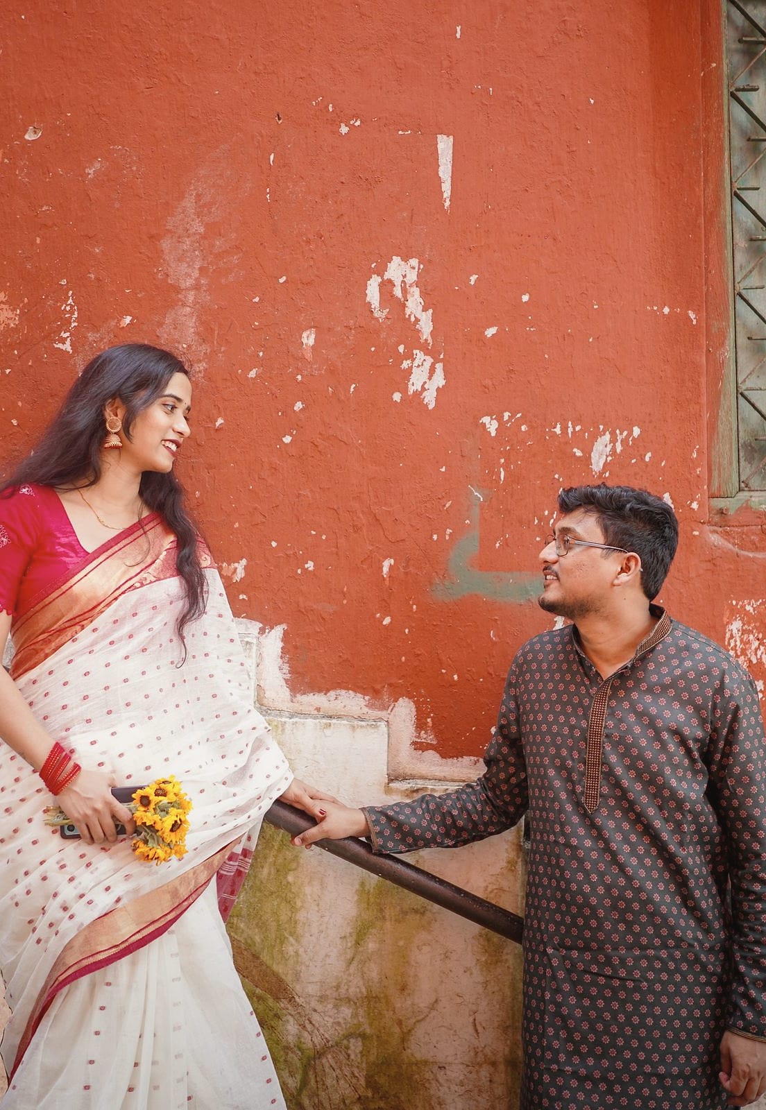 A couple on a stone staircase against a weathered red wall: she in a white saree with a red border, holding sunflowers, he in a dark kurta, looking at each other.