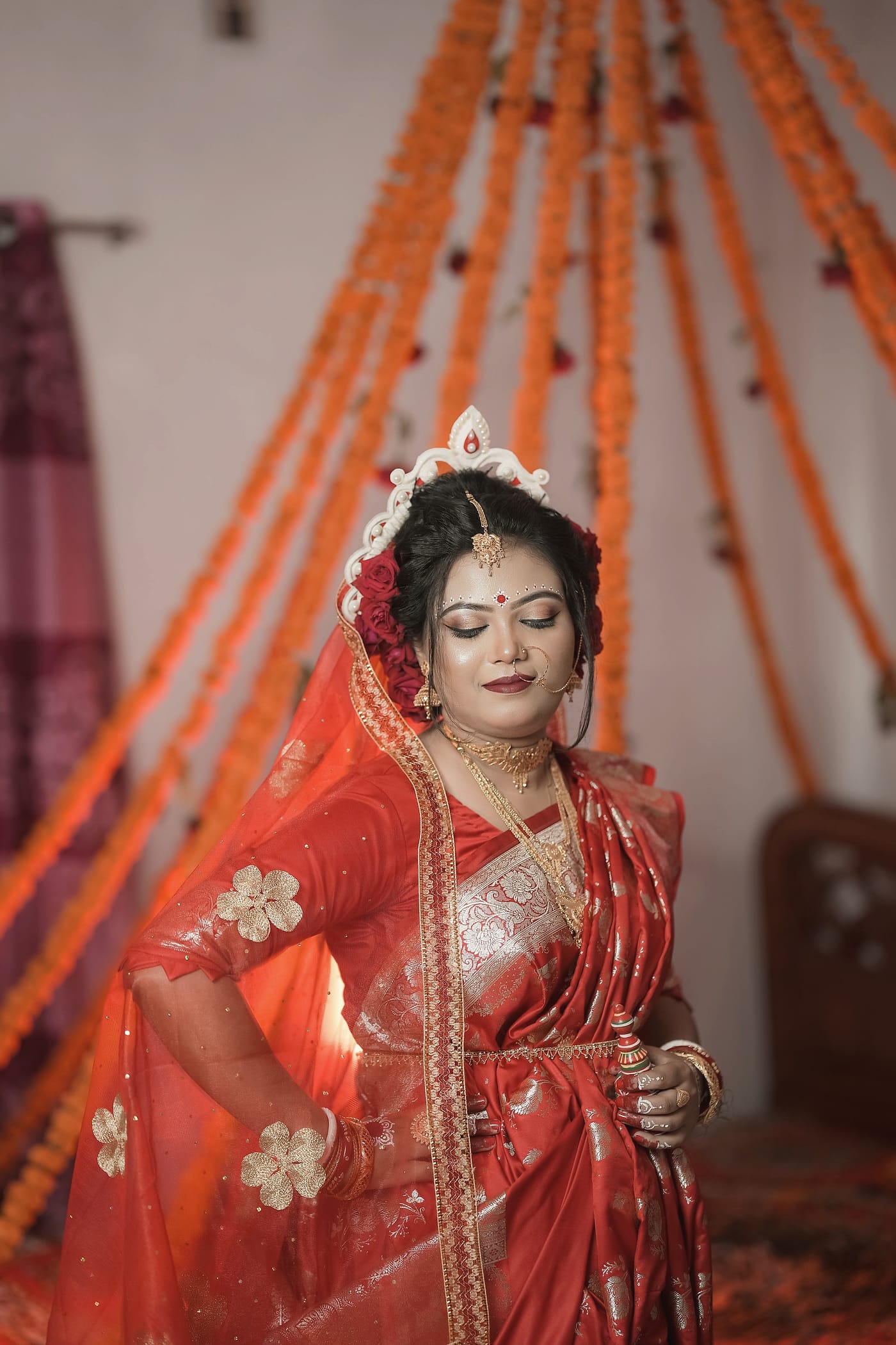 A bride in a red saree and white crown, one hand raised, against hanging marigold strings
