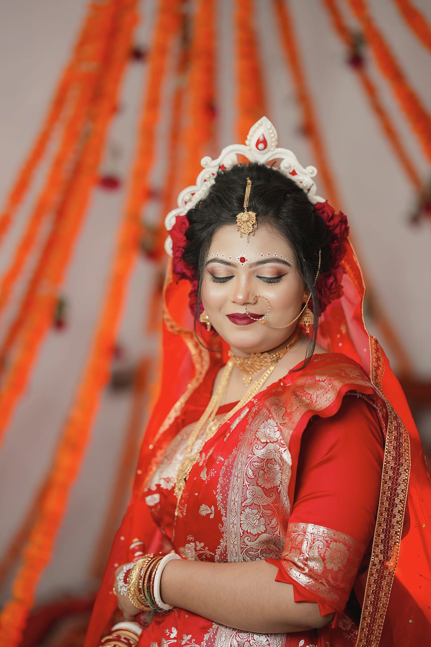 A bride in a crown and red saree, eyes lowered, hands together
