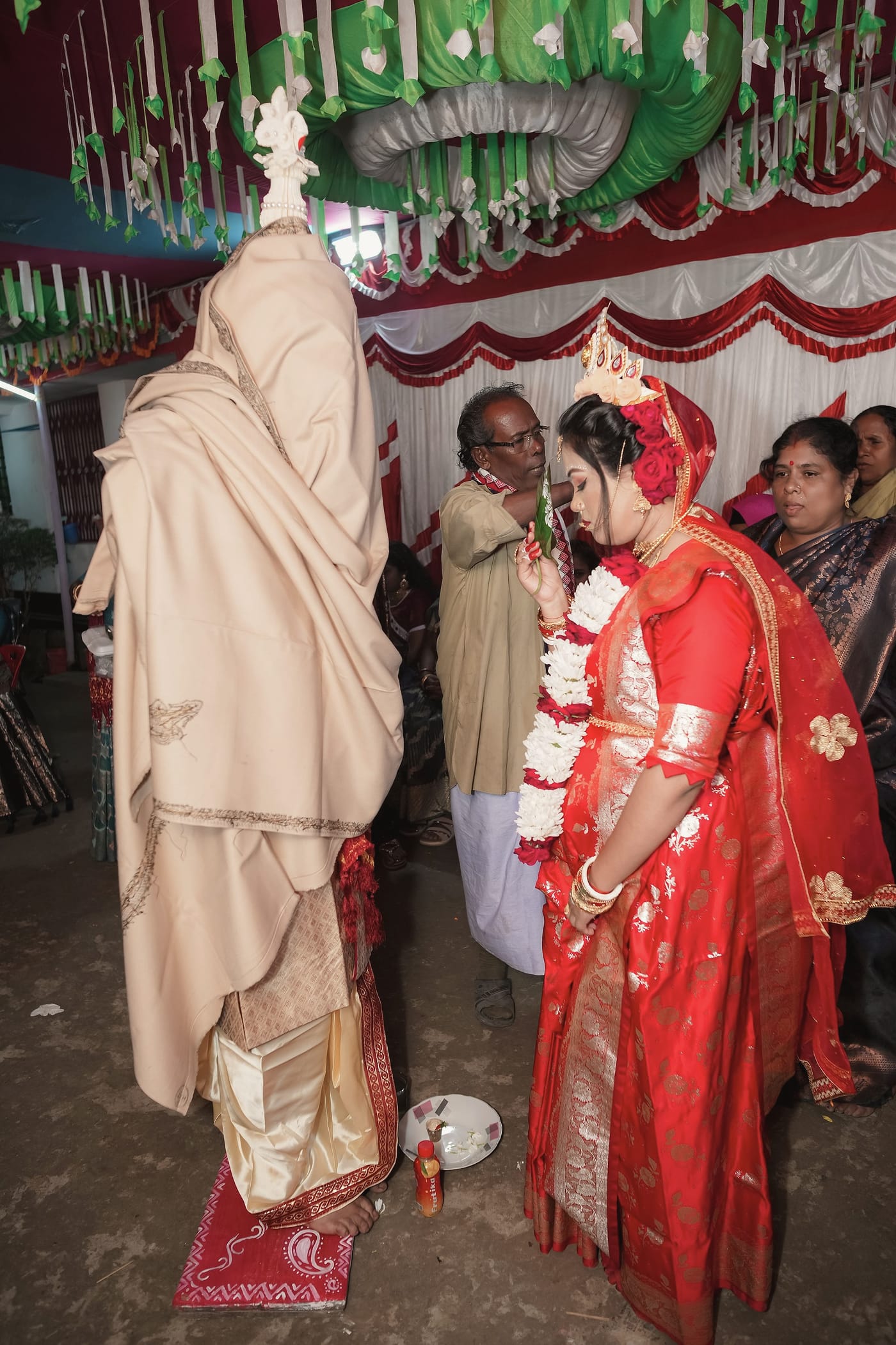 The groom standing in a cream shawl as the bride kneels beside him